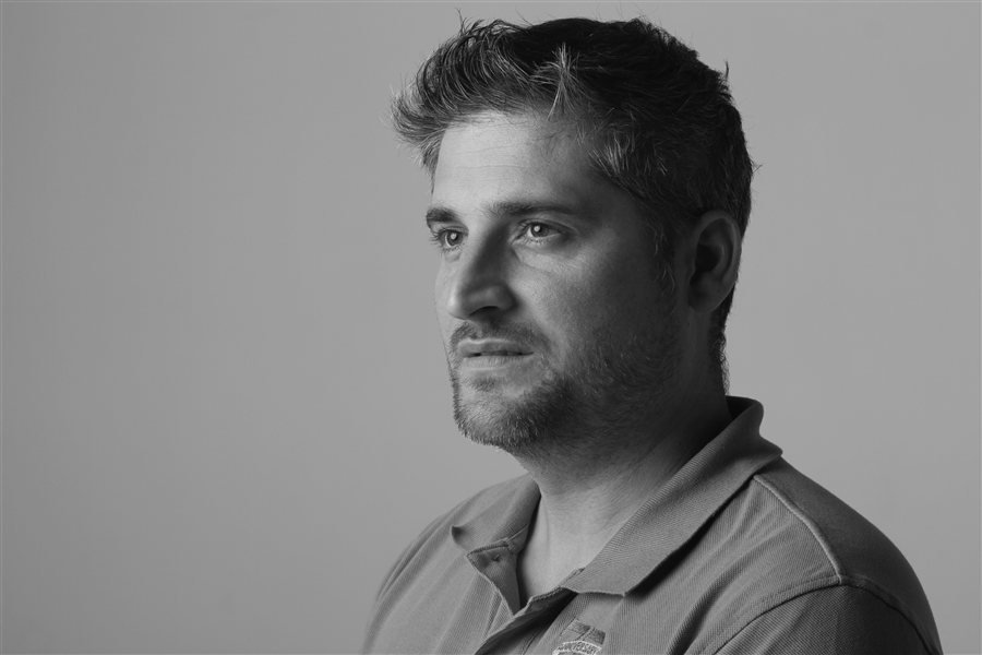 Black and white medium close-up portrait of a man with a beard, wearing a polo shirt, looking thoughtfully left.